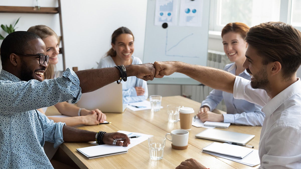 Diverse colleagues sitting at desk in boardroom feel happy and fist bumping greeting each other celebrating success at work